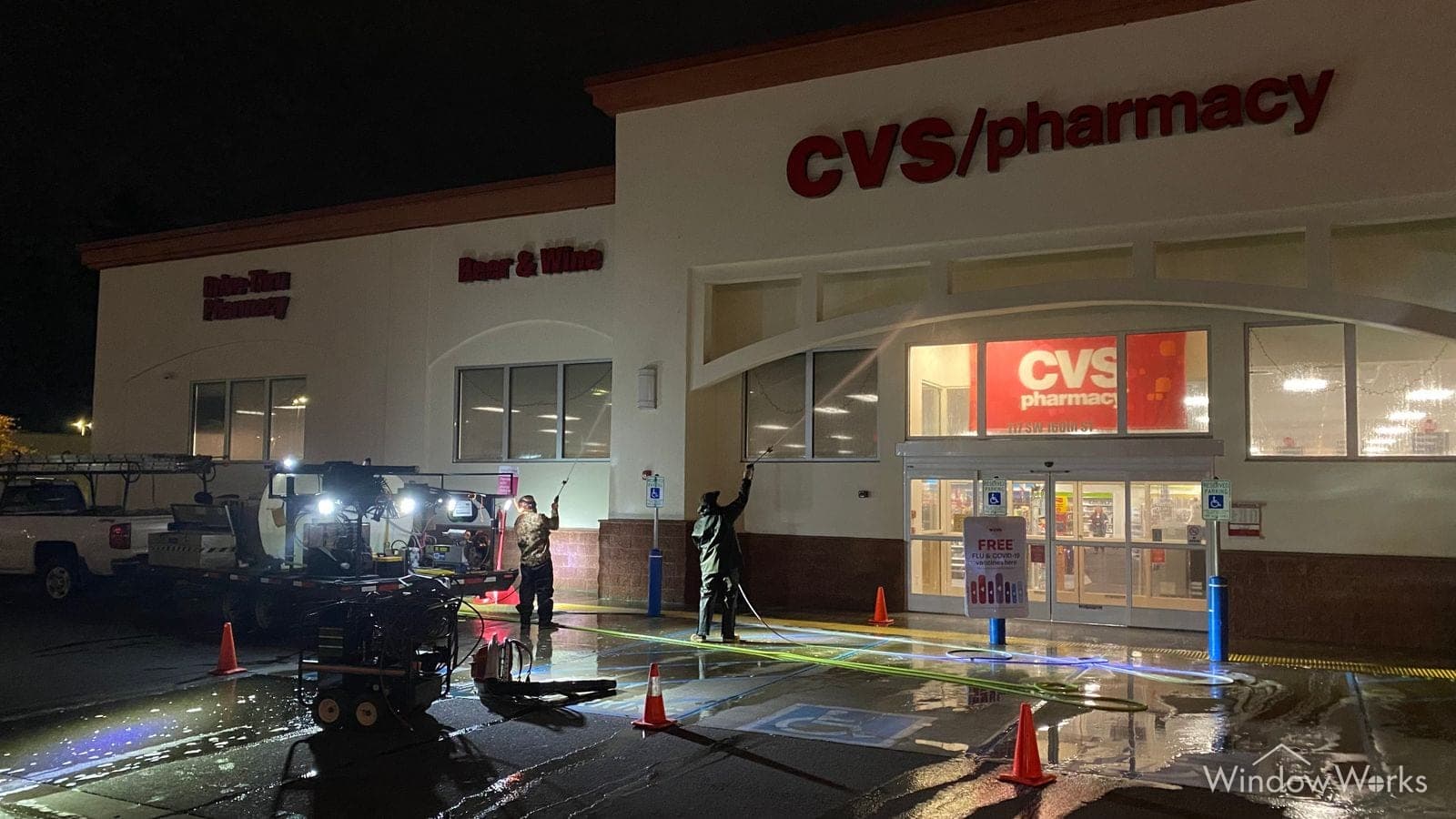 Two WindowWorks technicians washing a retail storefront overnight under work lights, cones and hot-water rig set up