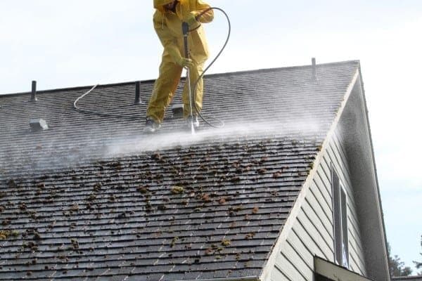 A worker in rain gear standing on a steep roof blasting moss with a high-pressure washer, spray flying off the shingles