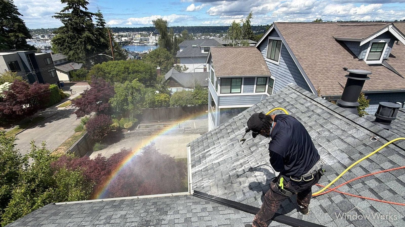 A WindowWorks technician soft washing a shingle roof above a Seattle neighborhood, a rainbow forming in the spray with the marina beyond