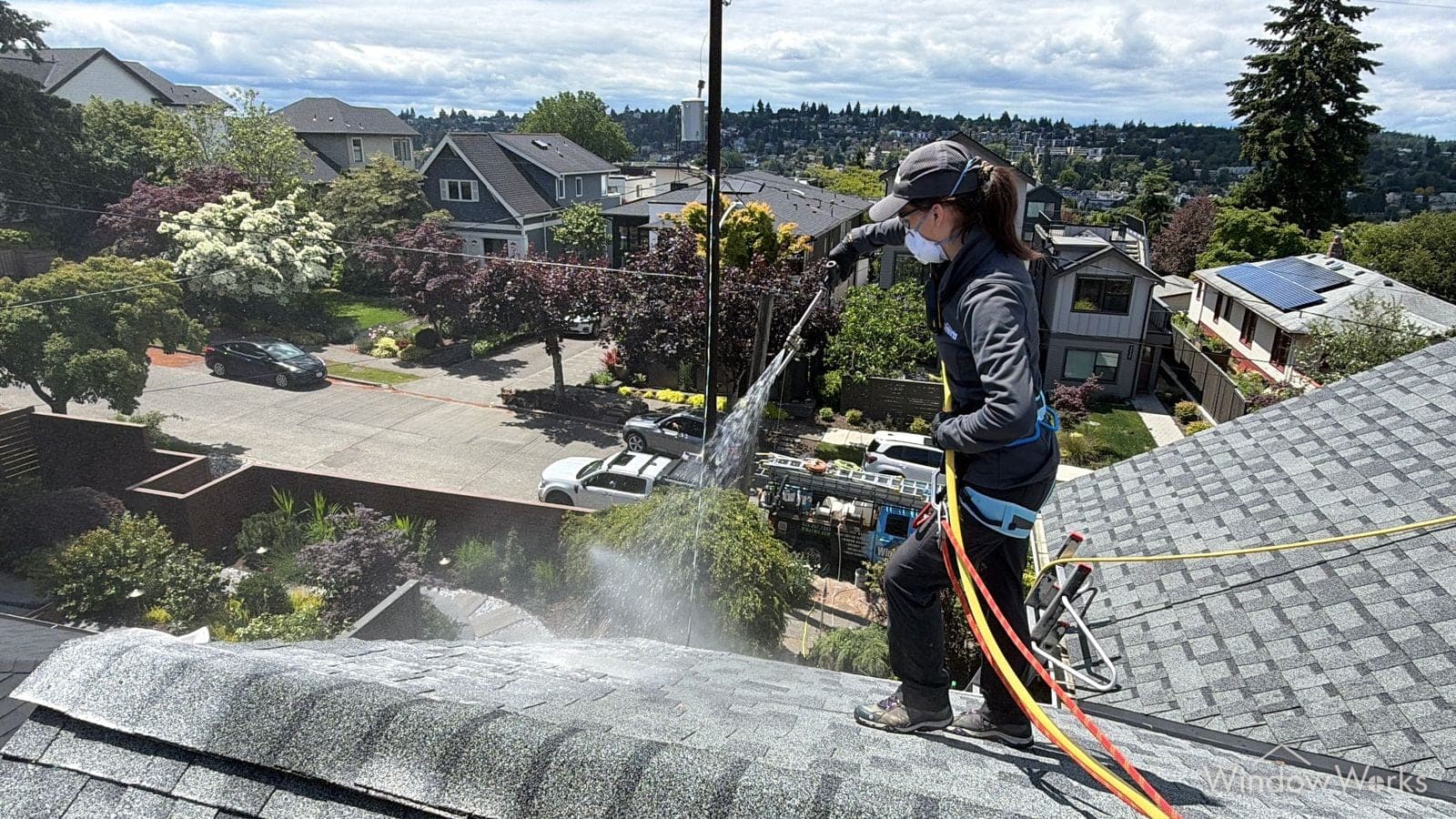 A WindowWorks technician on rope, soft washing a shingled roof high above a Seattle neighborhood on a sunny day