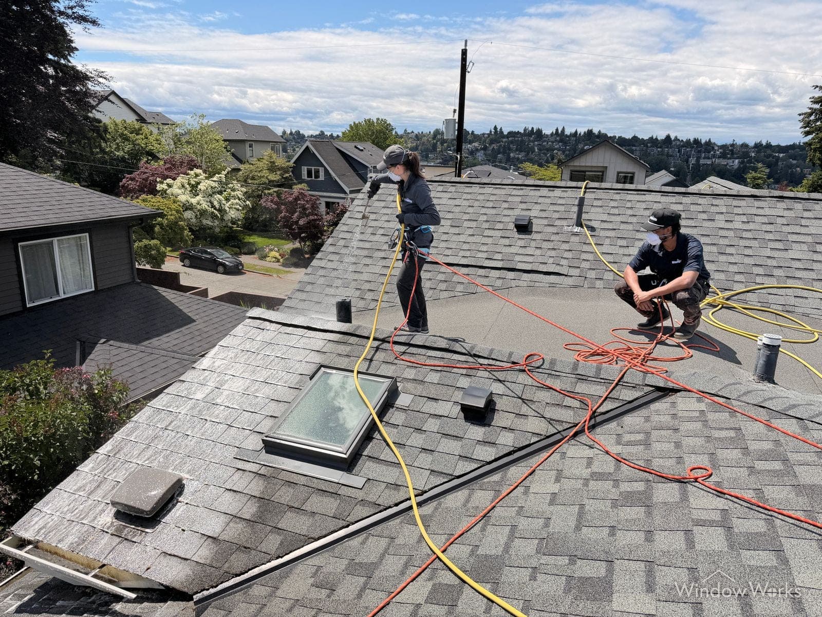 Two WindowWorks technicians soft washing a residential roof above a Seattle neighborhood, wearing harnesses and respirators