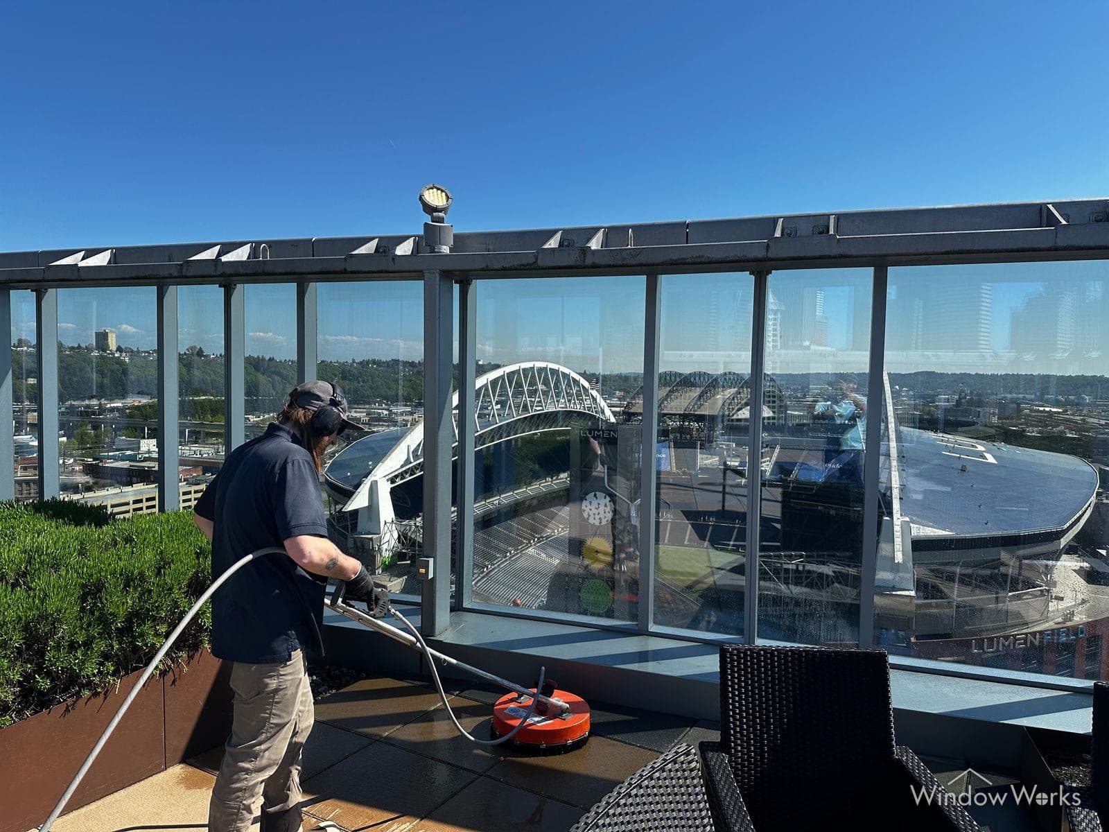 A WindowWorks technician surface-cleaning the pavers of a rooftop amenity deck with Lumen Field visible through the glass wind screens, Seattle.