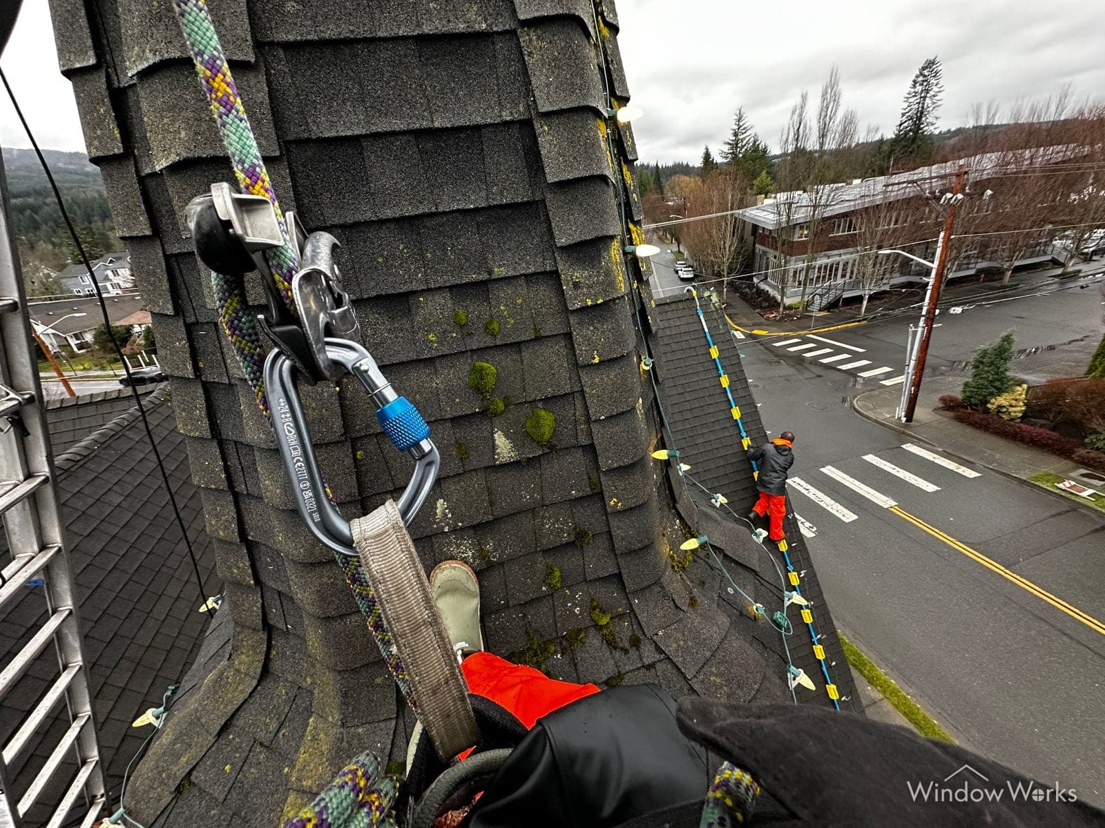 A WindowWorks technician's view from a steep shingled steeple, rope and carabiner anchored beside them, with a second roped technician working the roofline below and the street far underneath