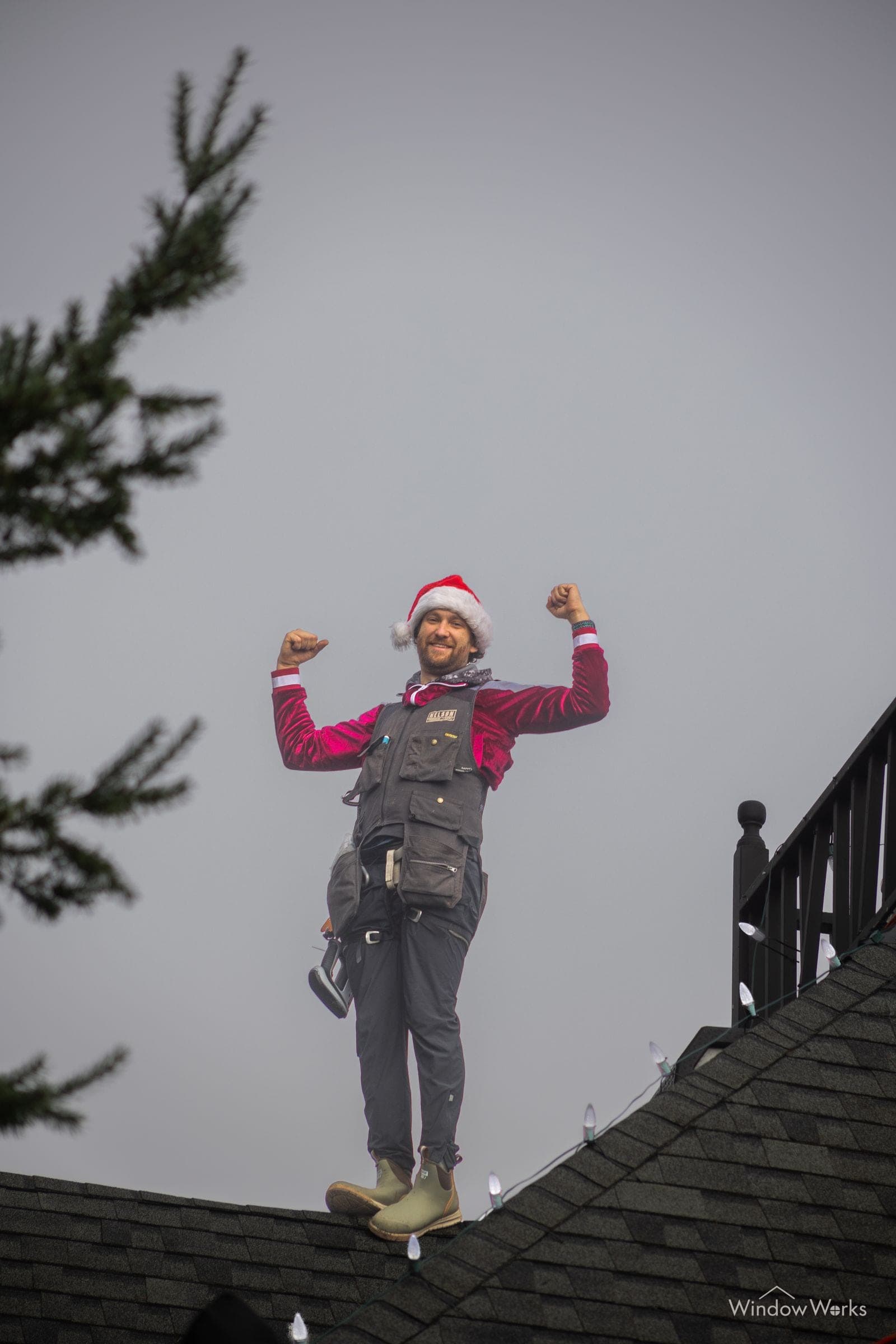 A WindowWorks technician in a Santa hat flexing both arms on a roofline after finishing a holiday light install