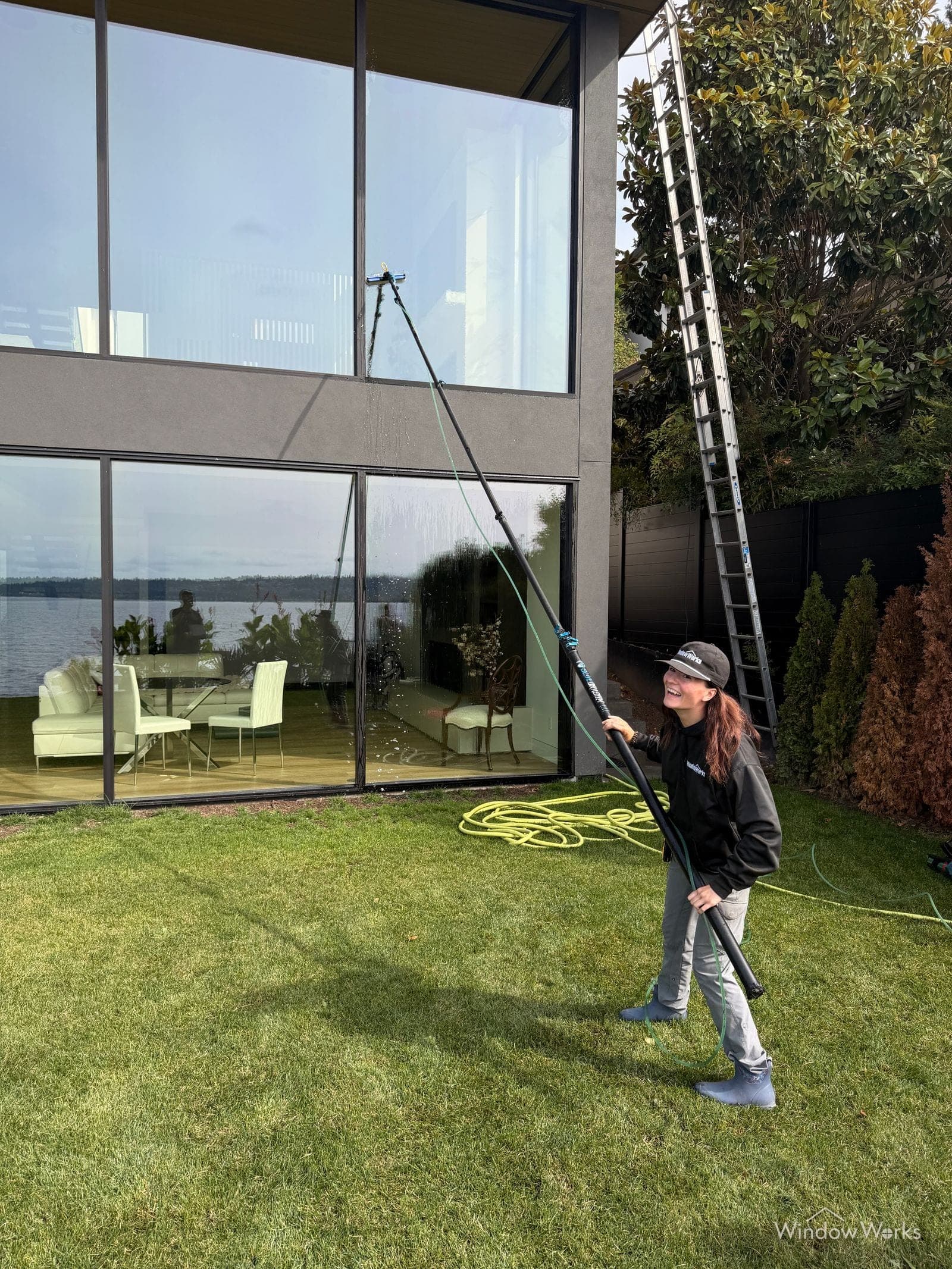 A smiling WindowWorks technician cleaning the tall upper windows of a lakefront home with a water fed pole, a long ladder against the wall nearby