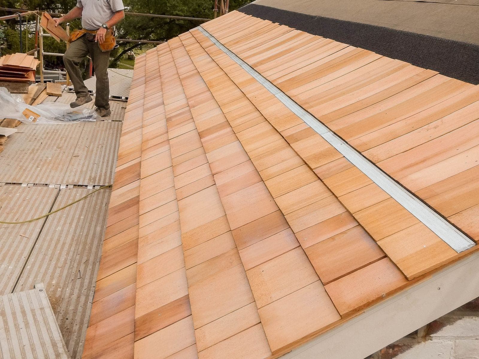 A bare zinc strip running along a new cedar shingle roof just below the ridge, with a roofer working nearby