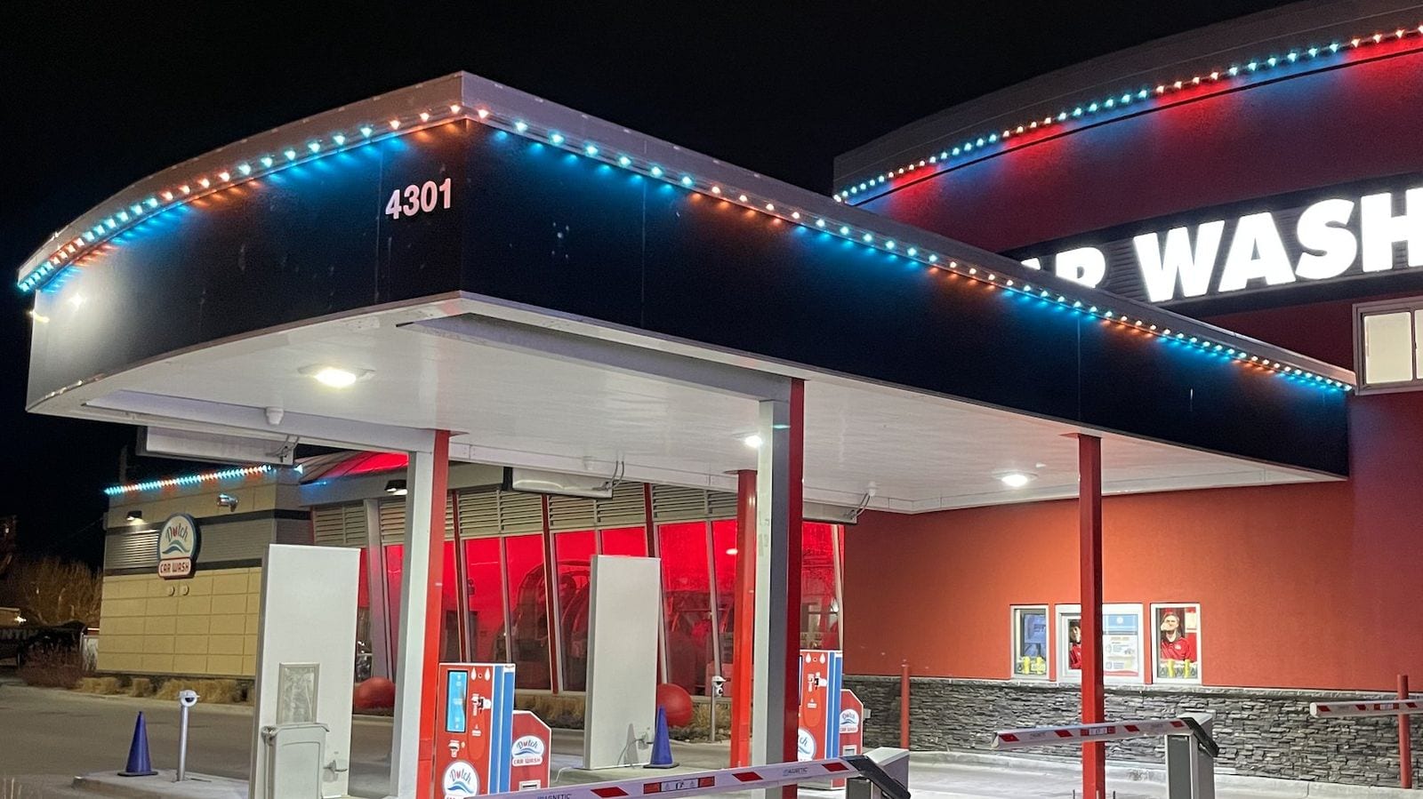 A car wash canopy at night with red, white and blue lights under the edge washing down the dark fascia in scallops